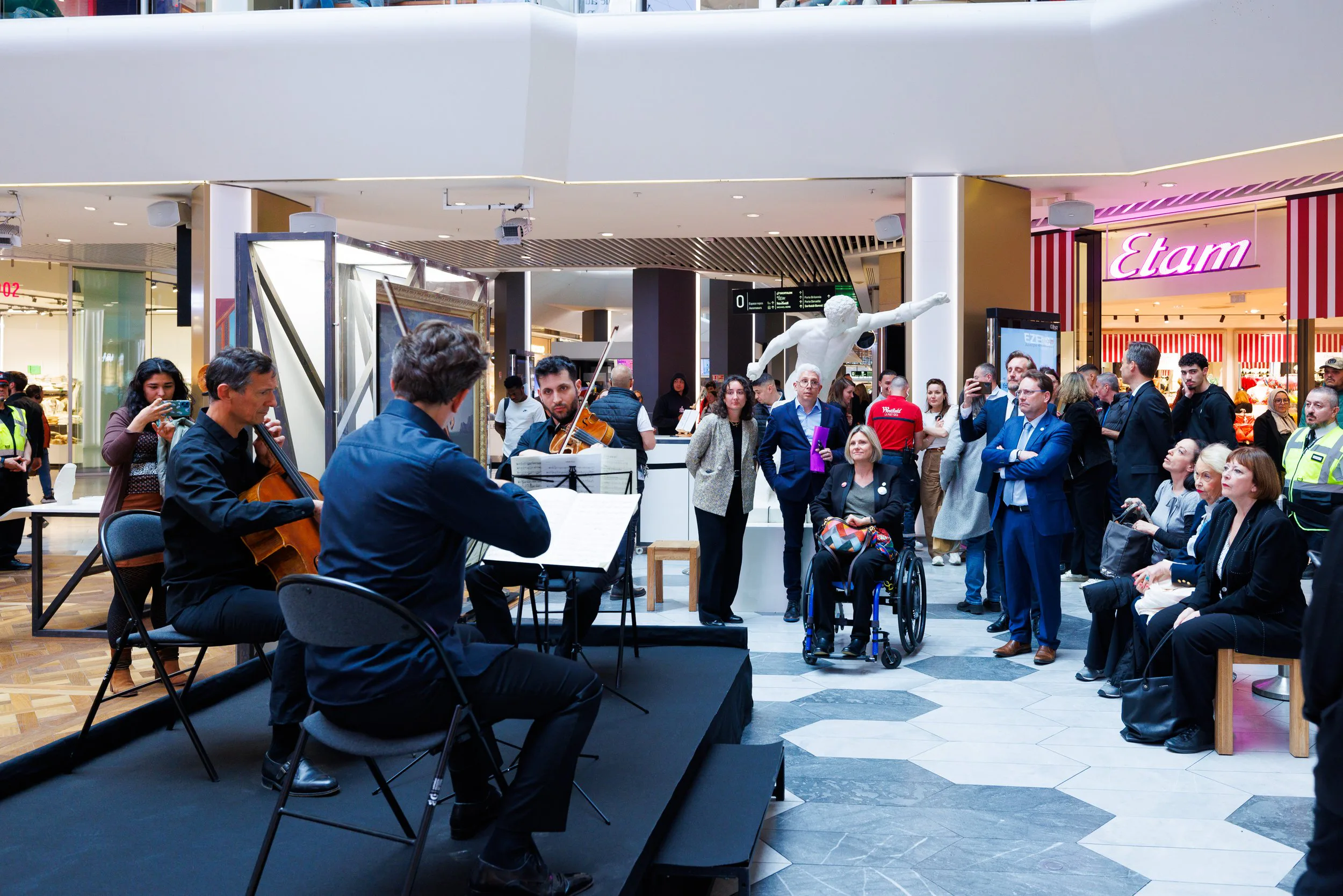 Un groupe de musiciens jouant du violon et du violoncelle dans un centre commercial, avec un public varié d'admirateurs, certains prenant des photos, et une sculpture blanche dans le fond, ainsi que plusieurs boutiques et enseignes visibles.