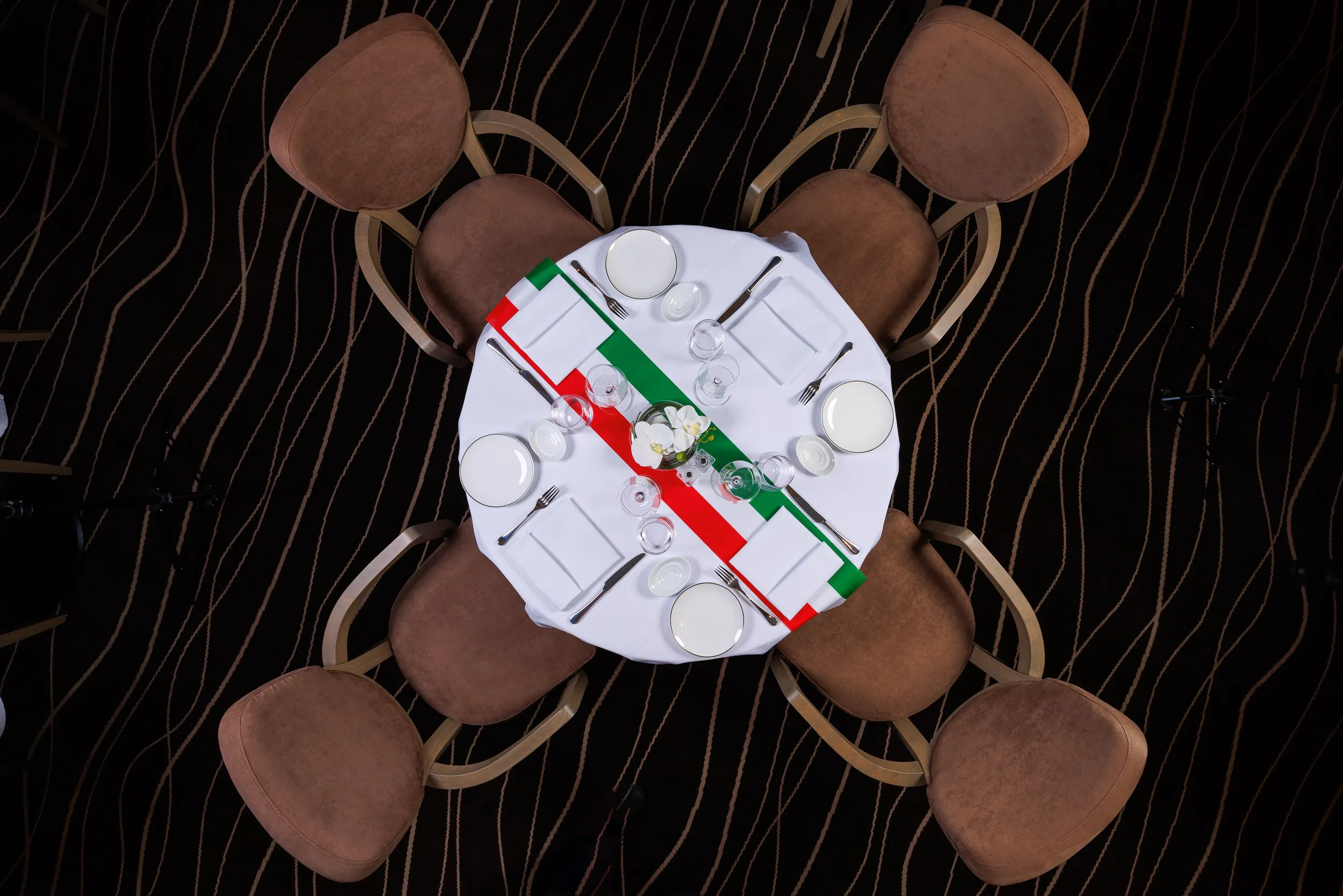 Table ronde dressée pour un repas, avec des assiettes, verres, fourchettes, couteaux, et nappes aux couleurs du drapeau italien, sur un tapis à motifs ondulés.