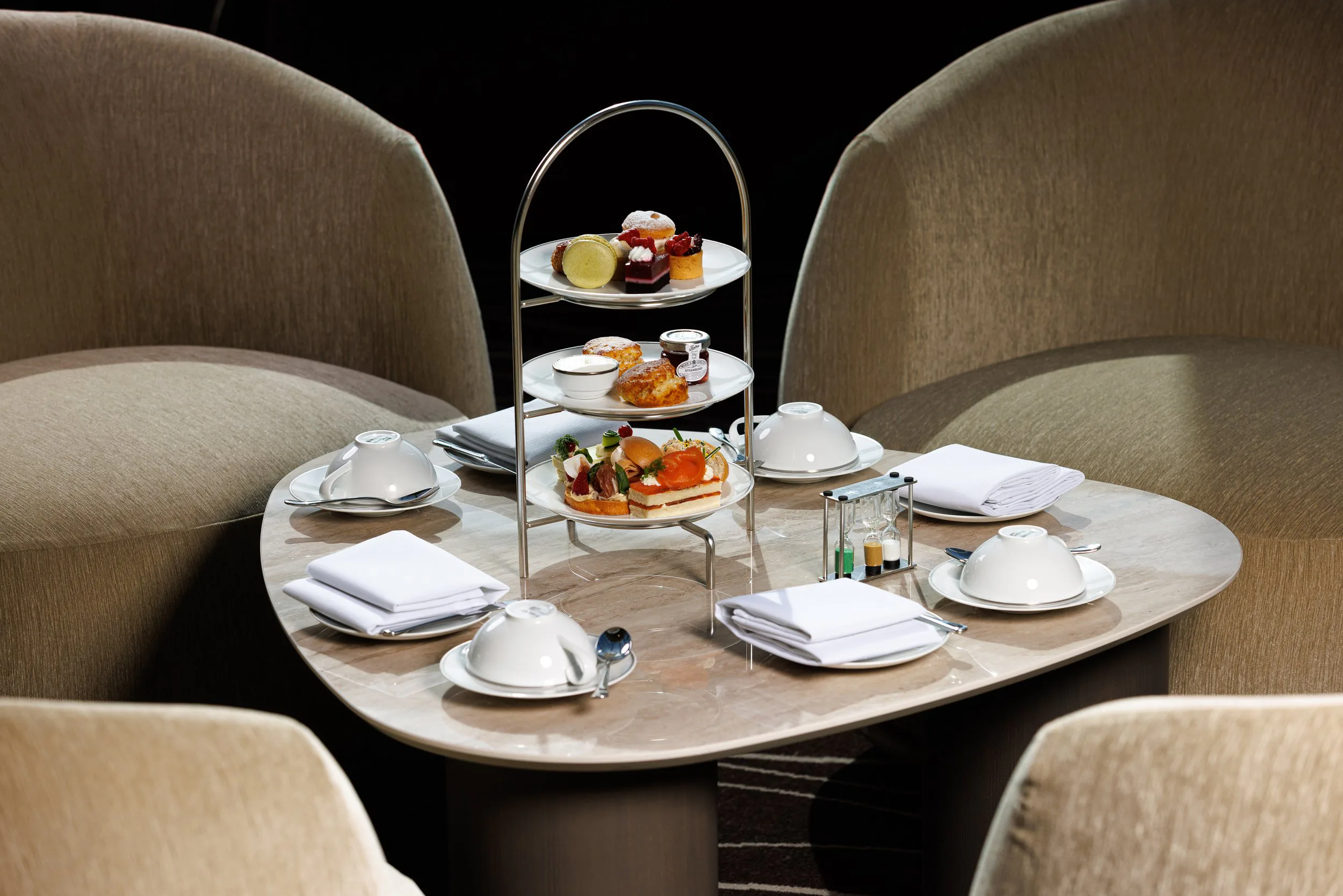 Table de petit déjeuner avec assiettes, couverts, serviettes et un plateau de desserts et sandwiches, entourée de chaises confortables.