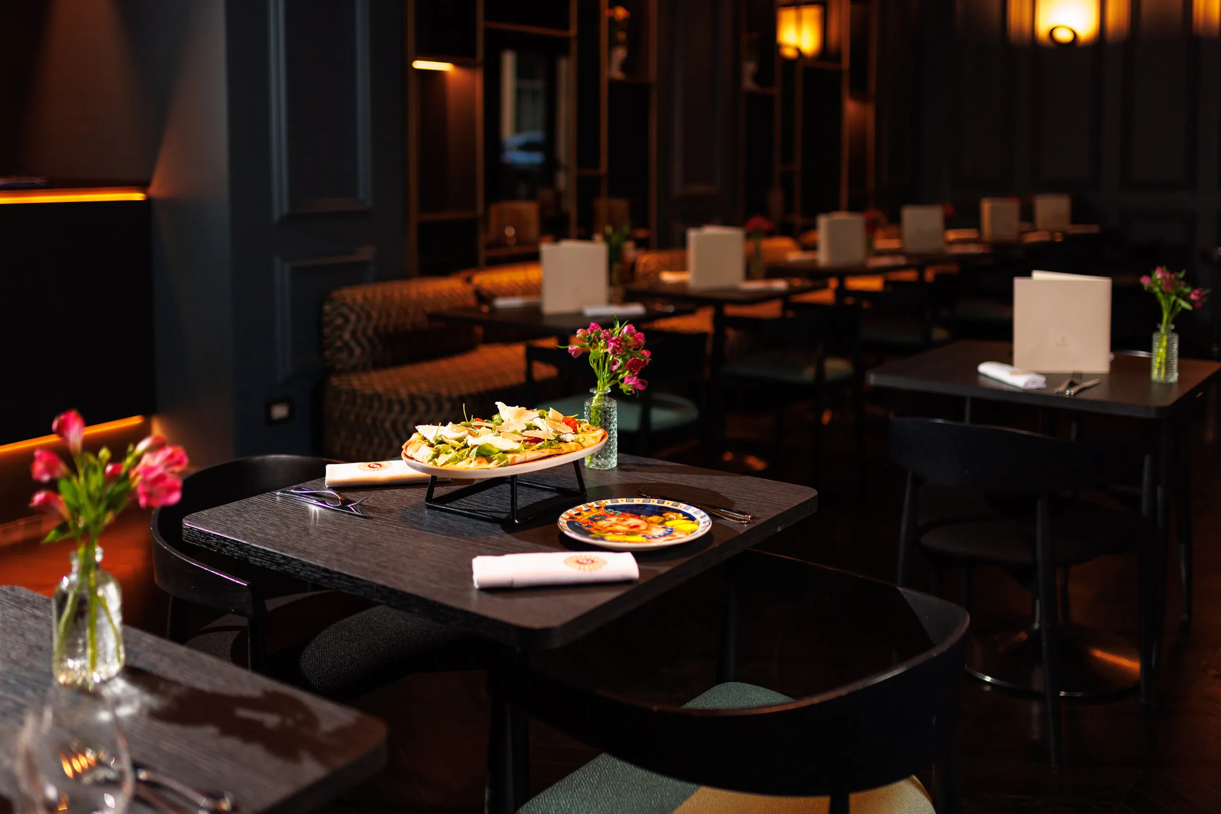 Une salle à manger d'un restaurant élégamment décorée avec des tables en bois noir, des fleurs roses en vases, et un plat de salade sur une table, dans une ambiance chaleureuse et intime.