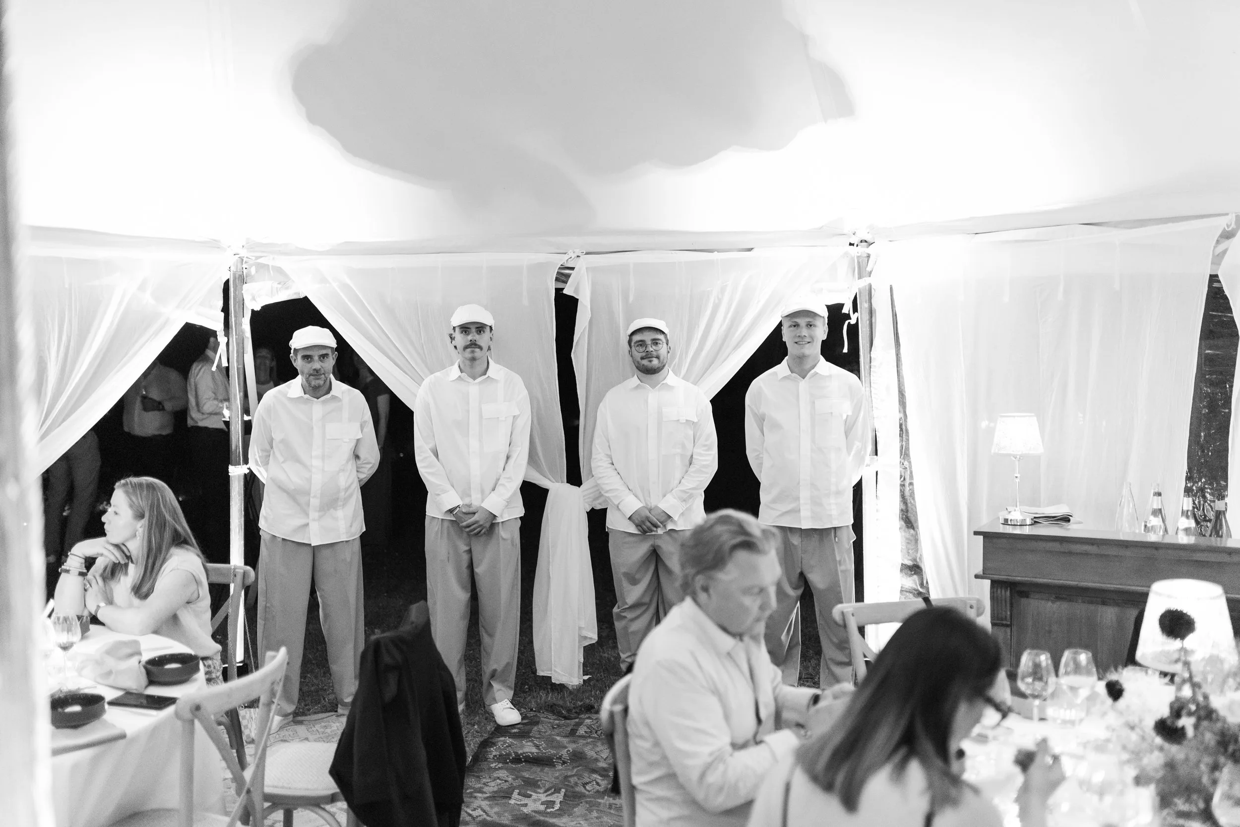 Quatre serveurs en uniforme blanc et chapeaux blancs se tiennent en rangée dans une tente lors d'un événement. Des invités sont assis à des tables en avant-plan, en train de dîner.