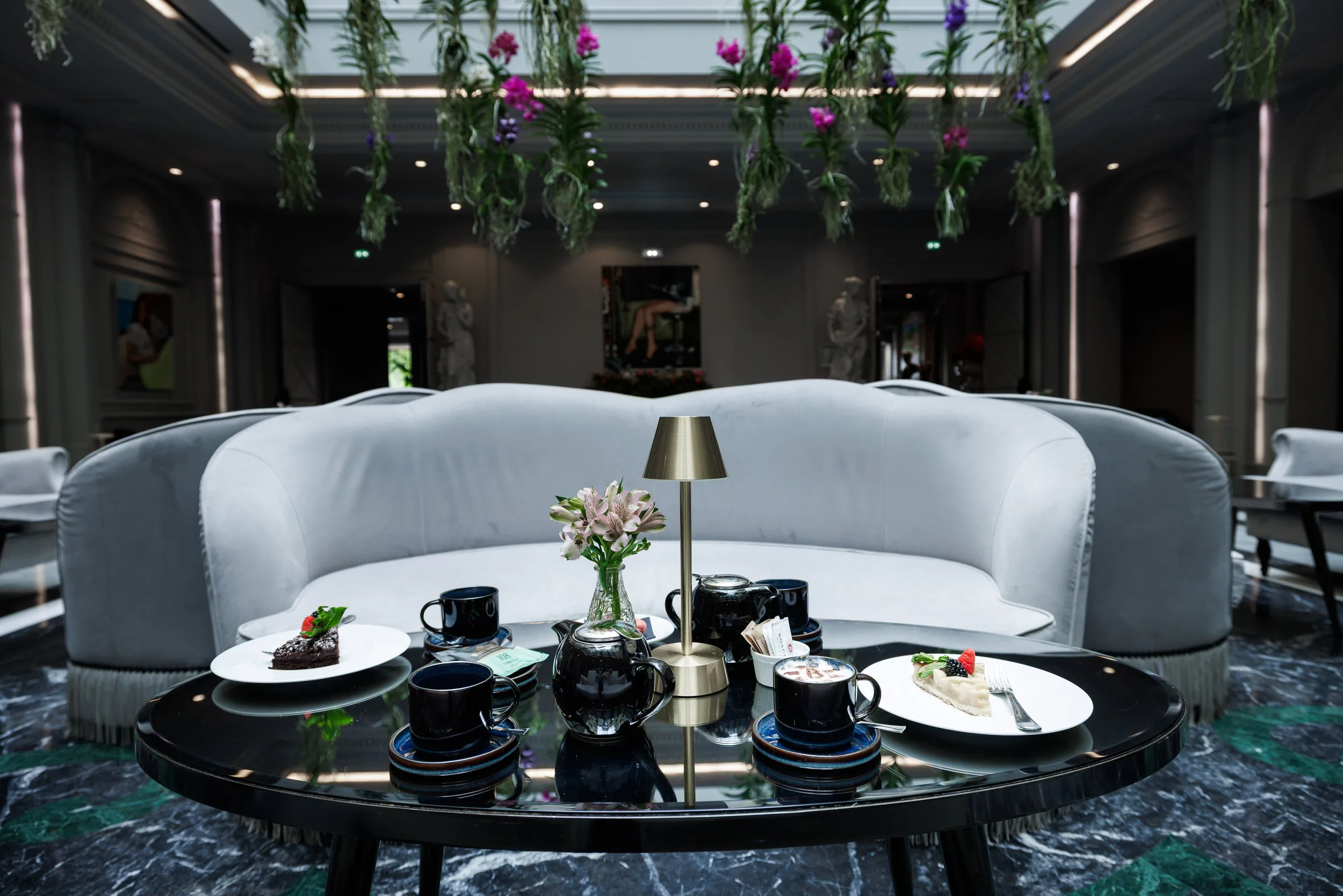 Salon lounge élégant avec un canapé blanc, une table ronde noire avec café, tasses, et gâteaux, décor floral suspendu au plafond, statues classiques en arrière-plan.