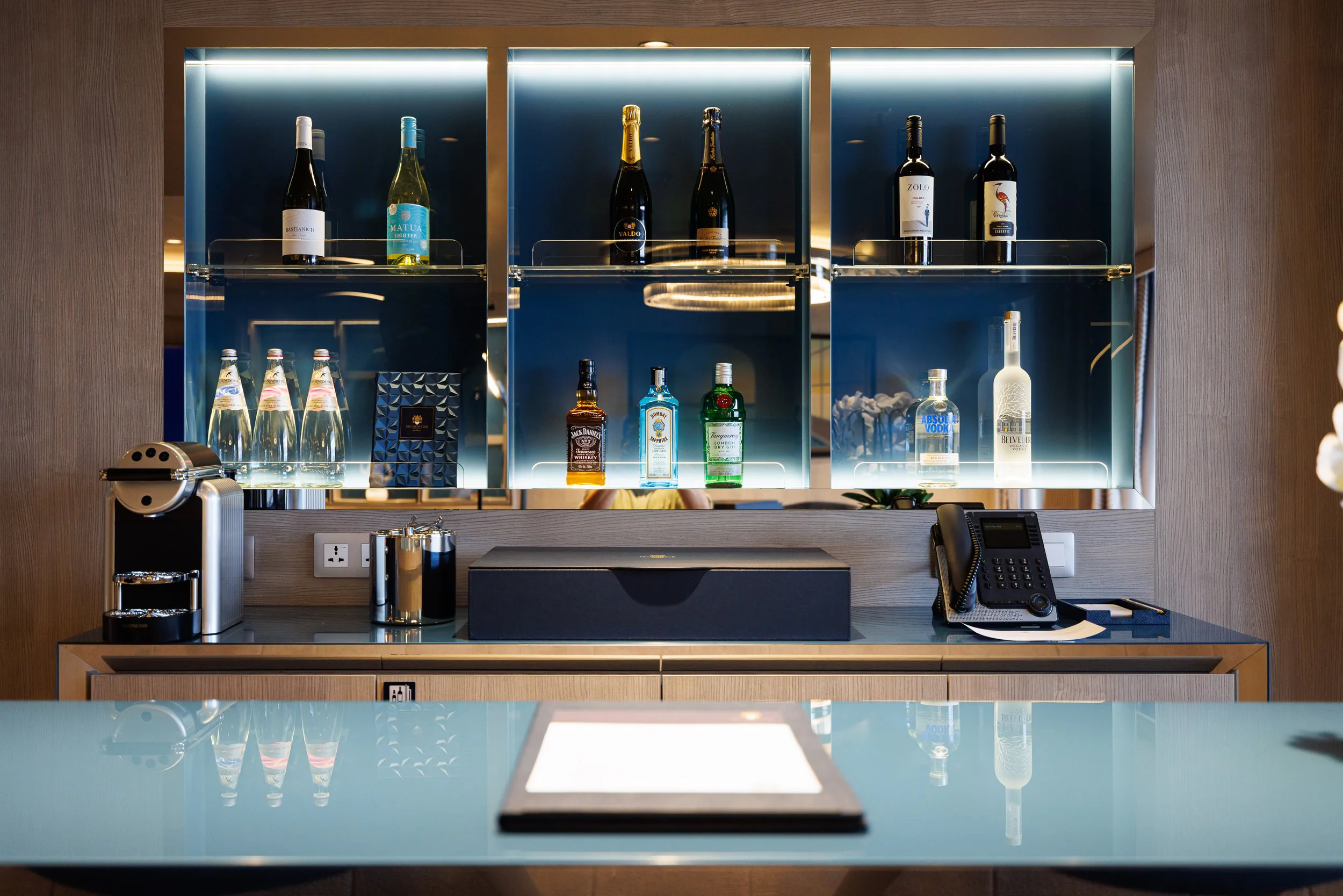 Espace de bar avec une étagère en verre contenant plusieurs bouteilles d'alcool et d'eau, un distributeur de café, un téléphone fixe et un clavier sur un comptoir en bois avec un fond en bois et un grand miroir.