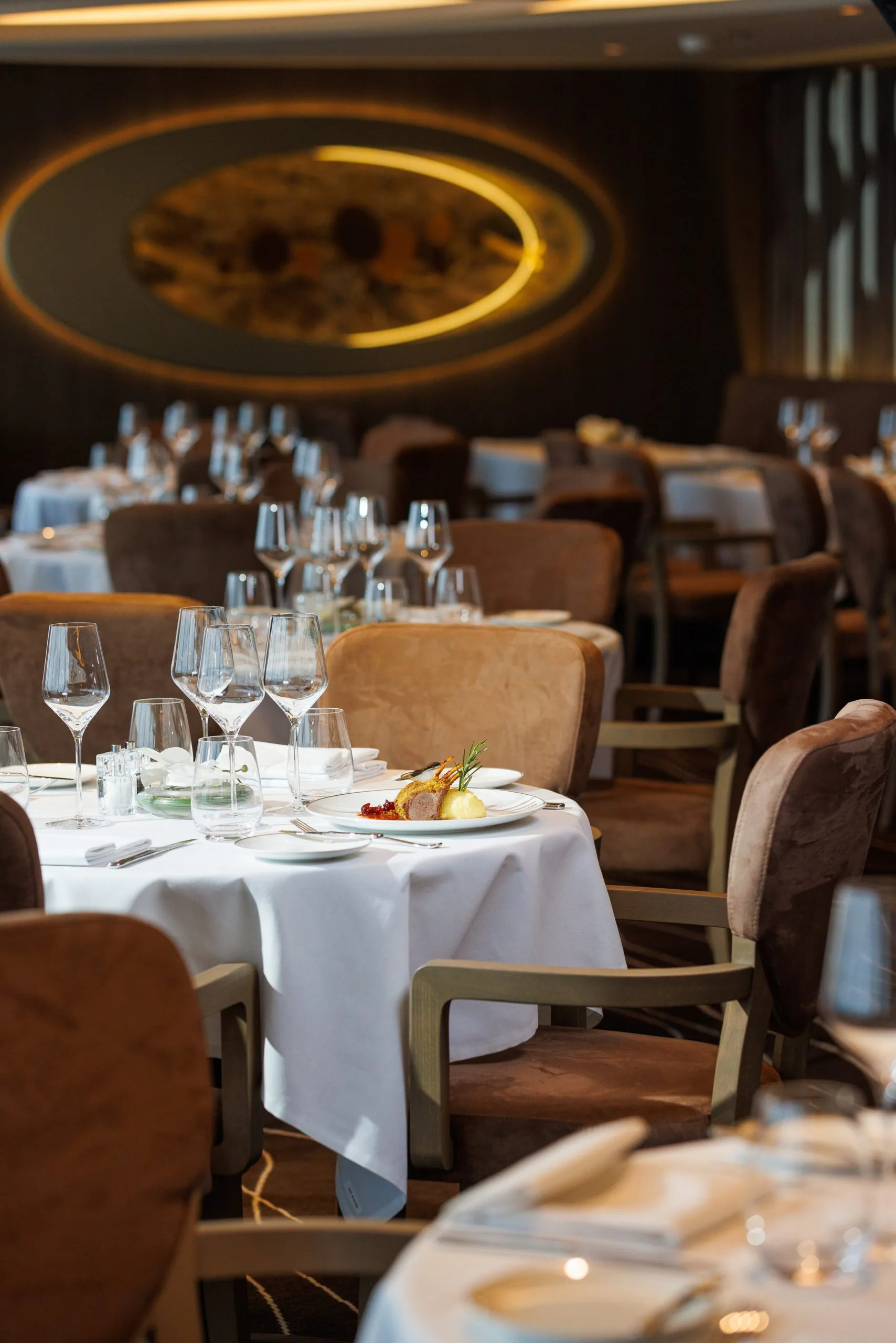 Salle à manger de restaurant élégante avec tables dressées, verres à vin, assiettes, et un plat de viande avec légumes, sous un éclairage chaleureux et un mur avec un grand tableau stylisé.