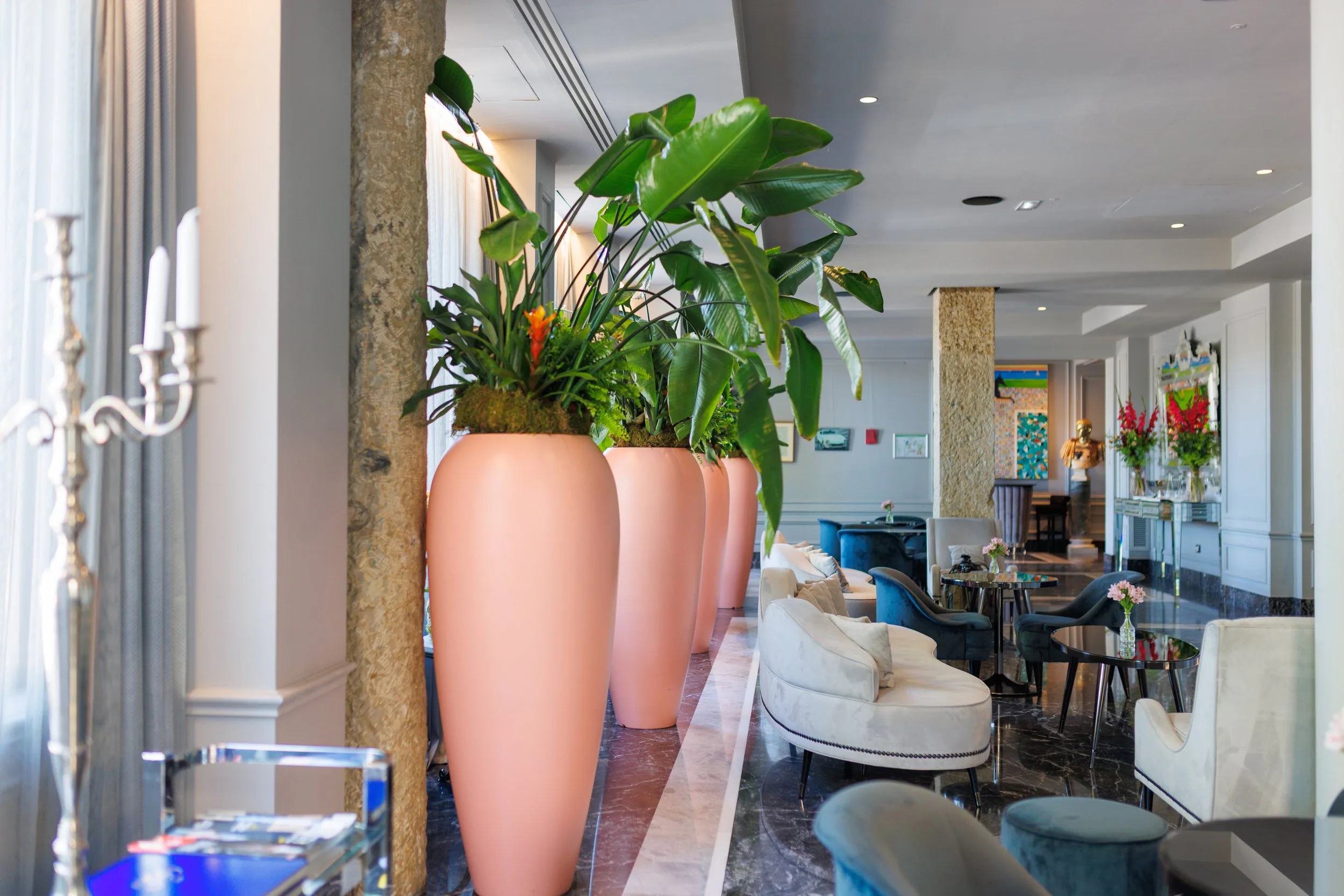 Salon élégant avec grandes plantes vertes dans de grands vases roses, fauteuils et petites tables ornées de fleurs, décor contemporain avec œuvres d'art sur les murs.