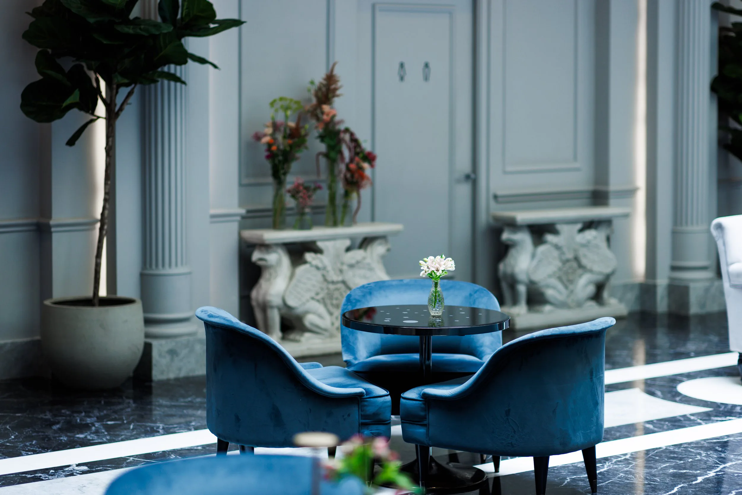Une petite table ronde en verre noir avec une vase contenant des fleurs blanches, entourée de quatre fauteuils bleus en velours dans un hall d'hôtel élégant avec des plantes et des décorations en marbre.