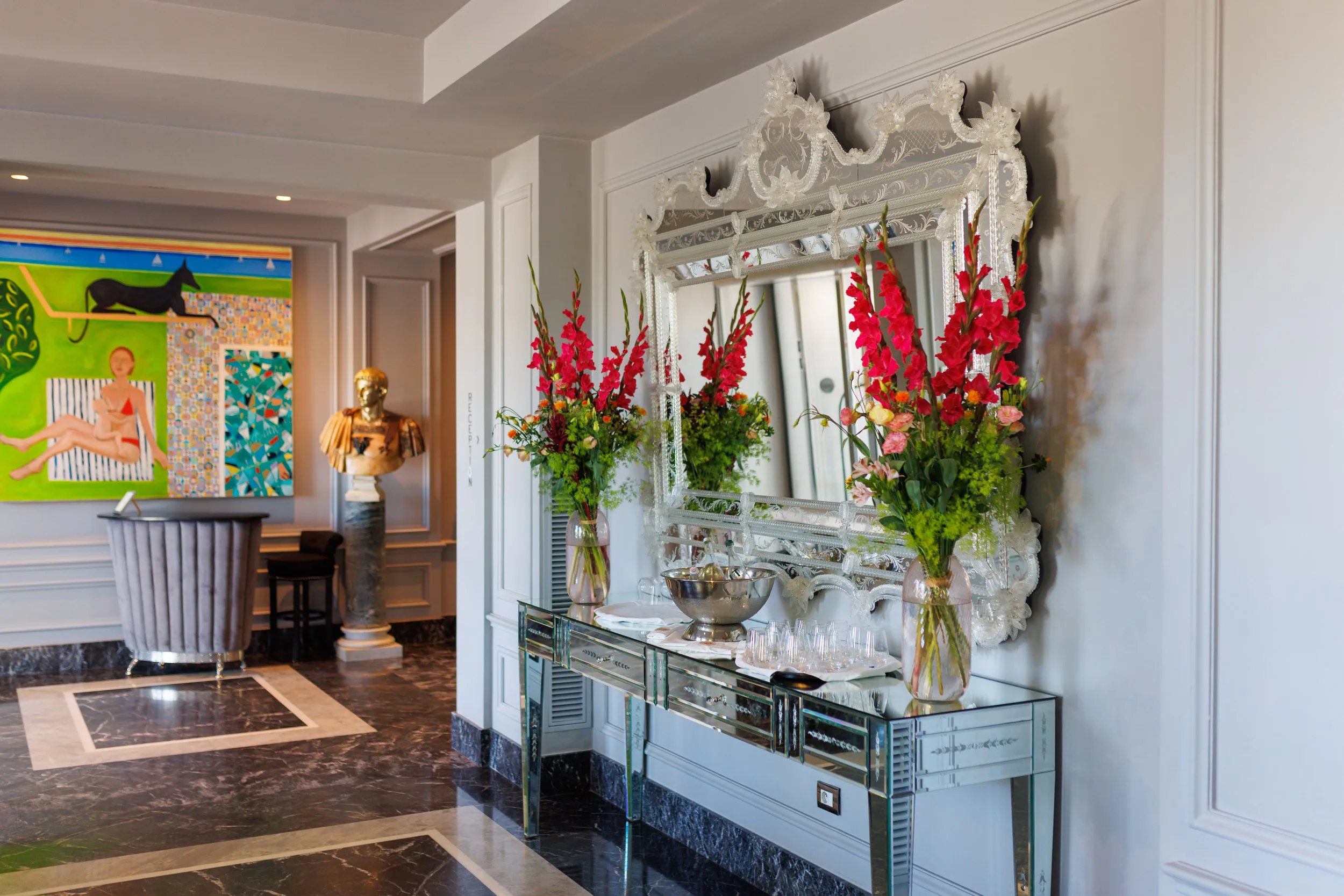 Intérieur élégant avec un grand miroir orné de détails sculptés, deux vases avec des fleurs roses et rouges sur une console en miroir, peinture colorée dans la salle voisine, statue en or beige et mur blanc decoré