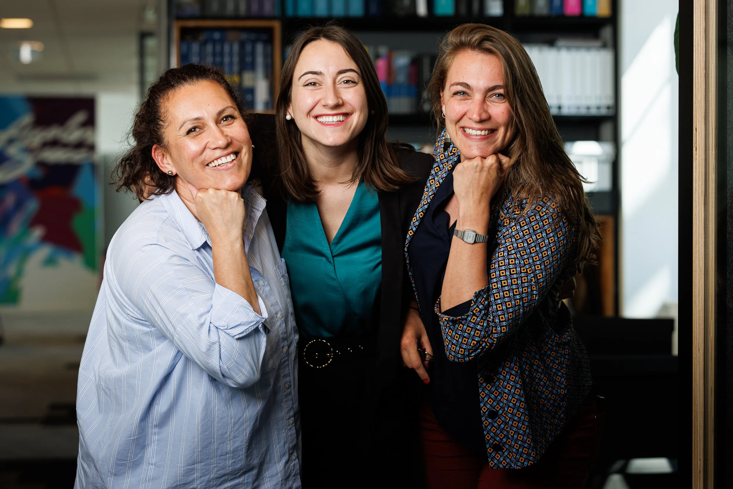 Trois femmes souriantes posant ensemble en intérieur, arrière-plan avec bibliothèque et peinture