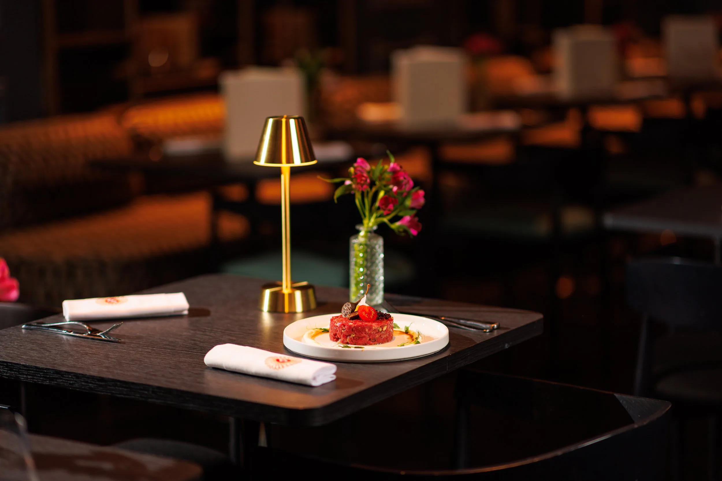 Table de restaurant élégante avec un plat de viande, un vase de fleurs roses, une lampe dorée et des couverts, fond flou.
