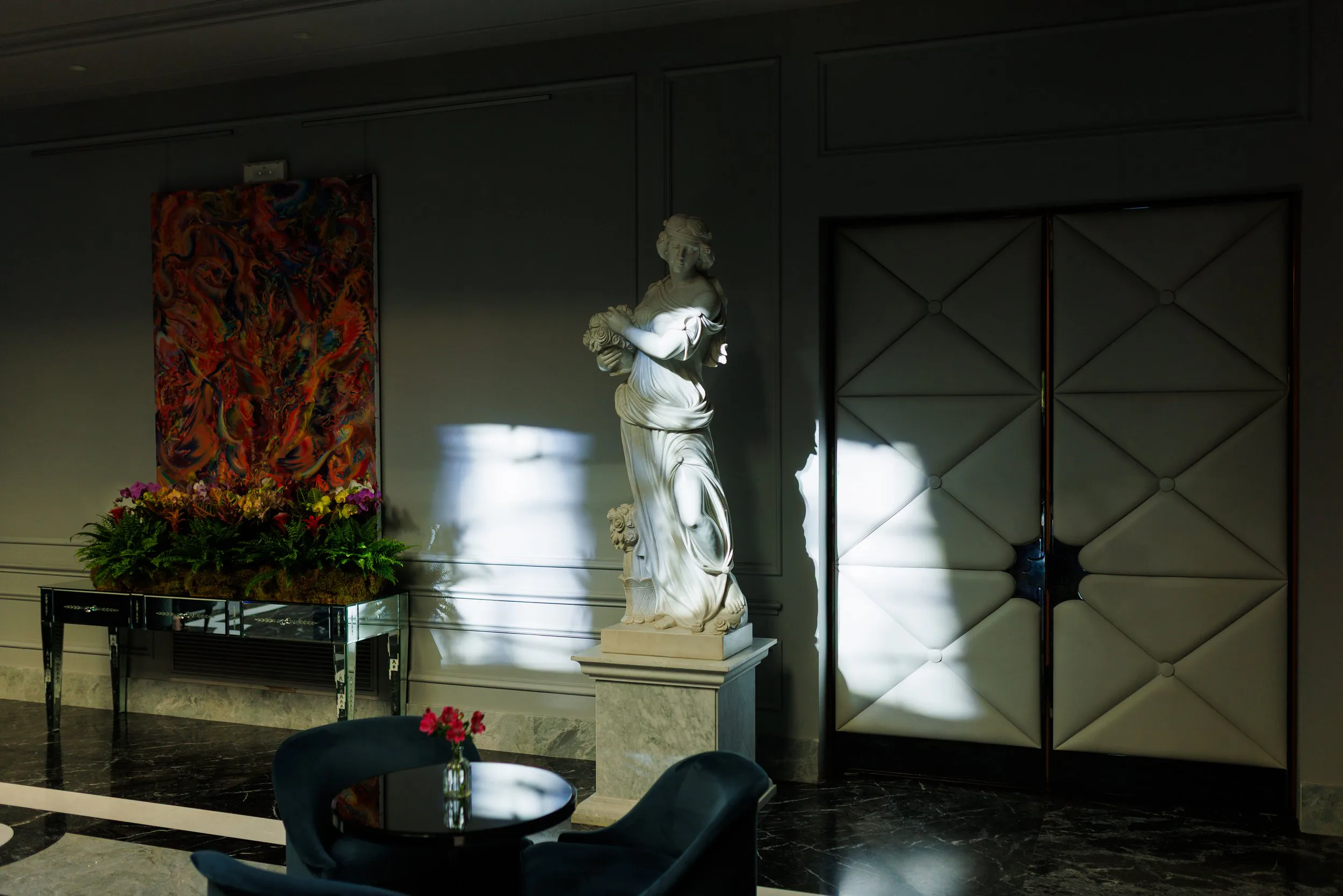 Une sculpture en marbre d'une femme tenant un bouquet, située dans un hall sombre avec des lumières créant des ombres sur le mur, un tableau coloré et un coin avec une table et des chaises avec un vase de fleurs.