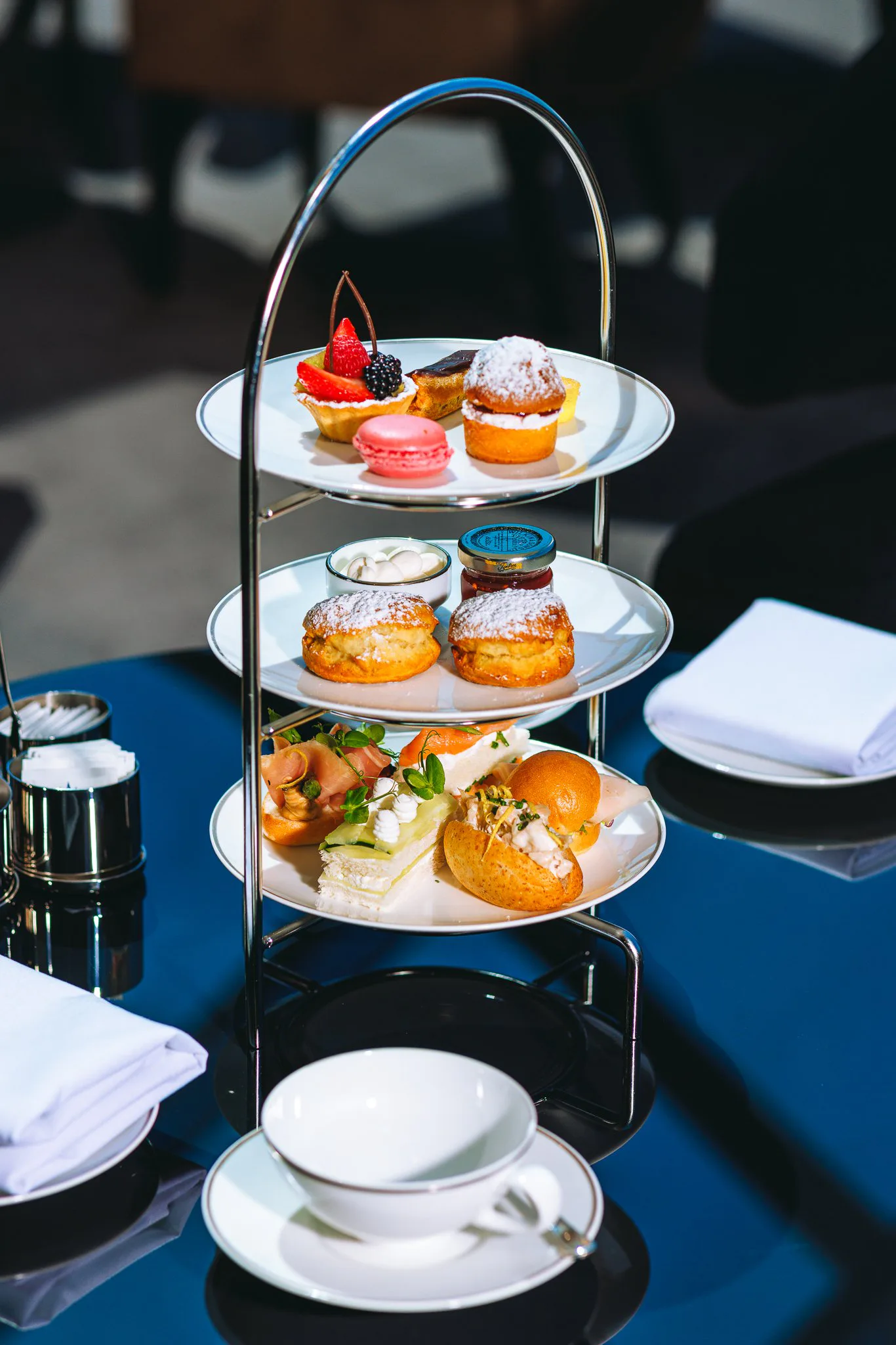 Plateau de goûter avec trois étages, comprenant divers desserts et amuse-bouches, sur une table avec assiettes, tasses, et couverts.