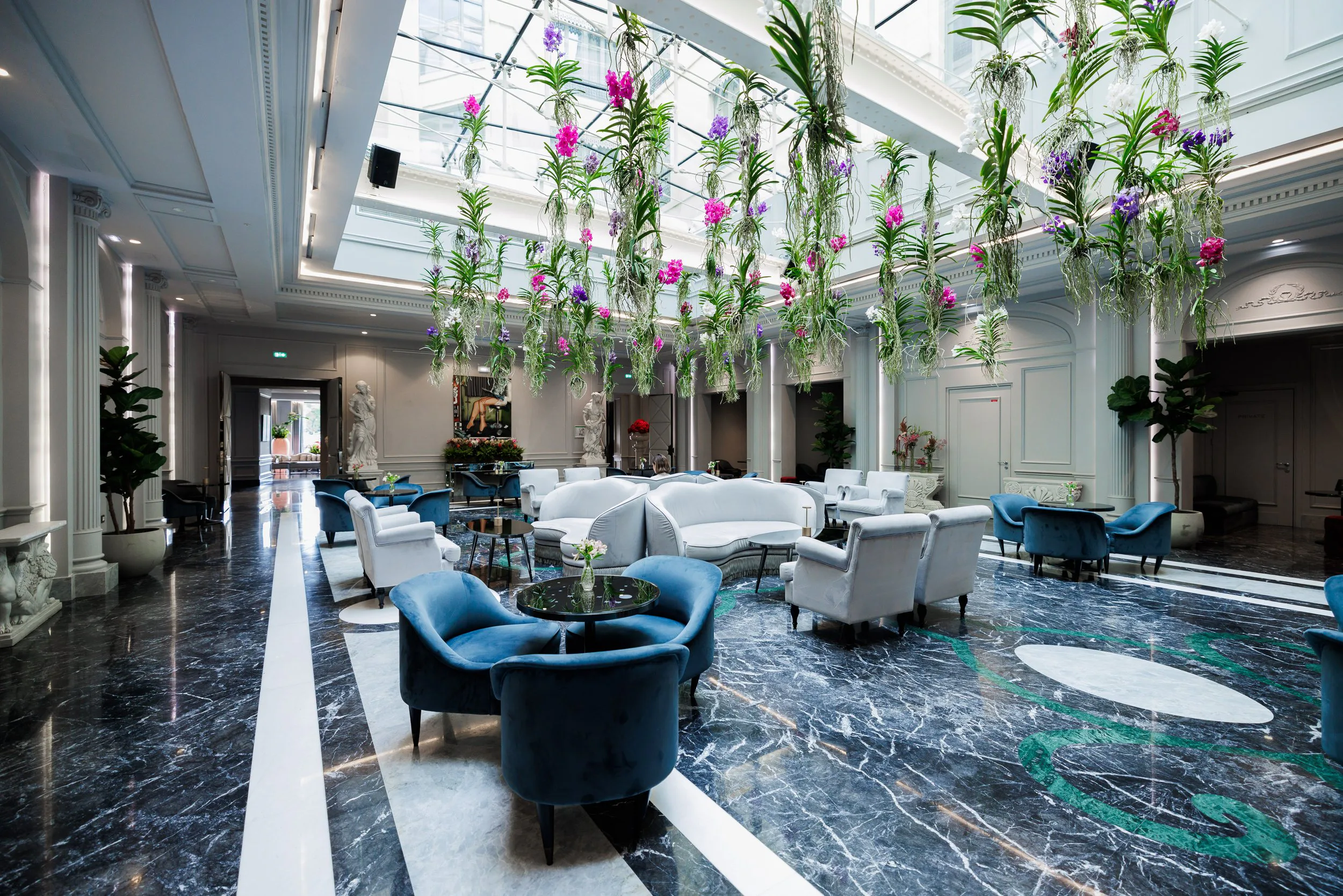 Un grand salon élégant avec des fauteuils et canapés blancs et bleus, des tables basses en marbre noir, et un plafond en verre avec des plantes suspendues colorées. Murs blancs avec sculptures et œuvres d'art, sol en marbre noir avec des motifs verts