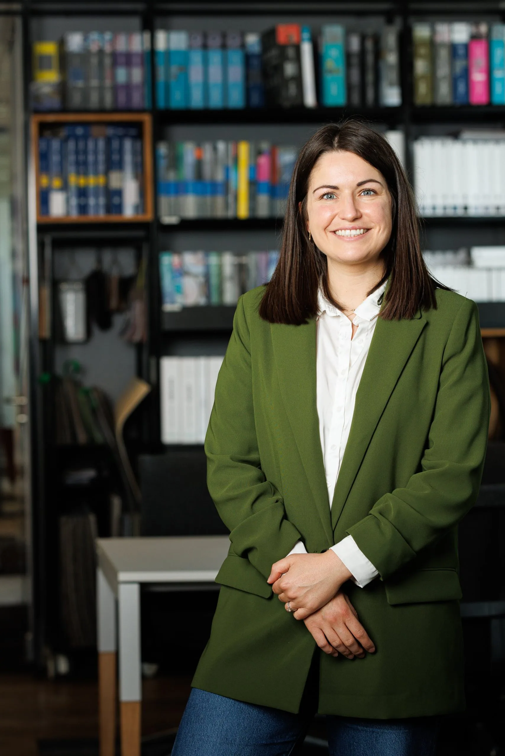 Une femme souriante portant un blazer vert et une chemise blanche, se tenant dans un bureau avec des étagères pleines de livres en arrière-plan.