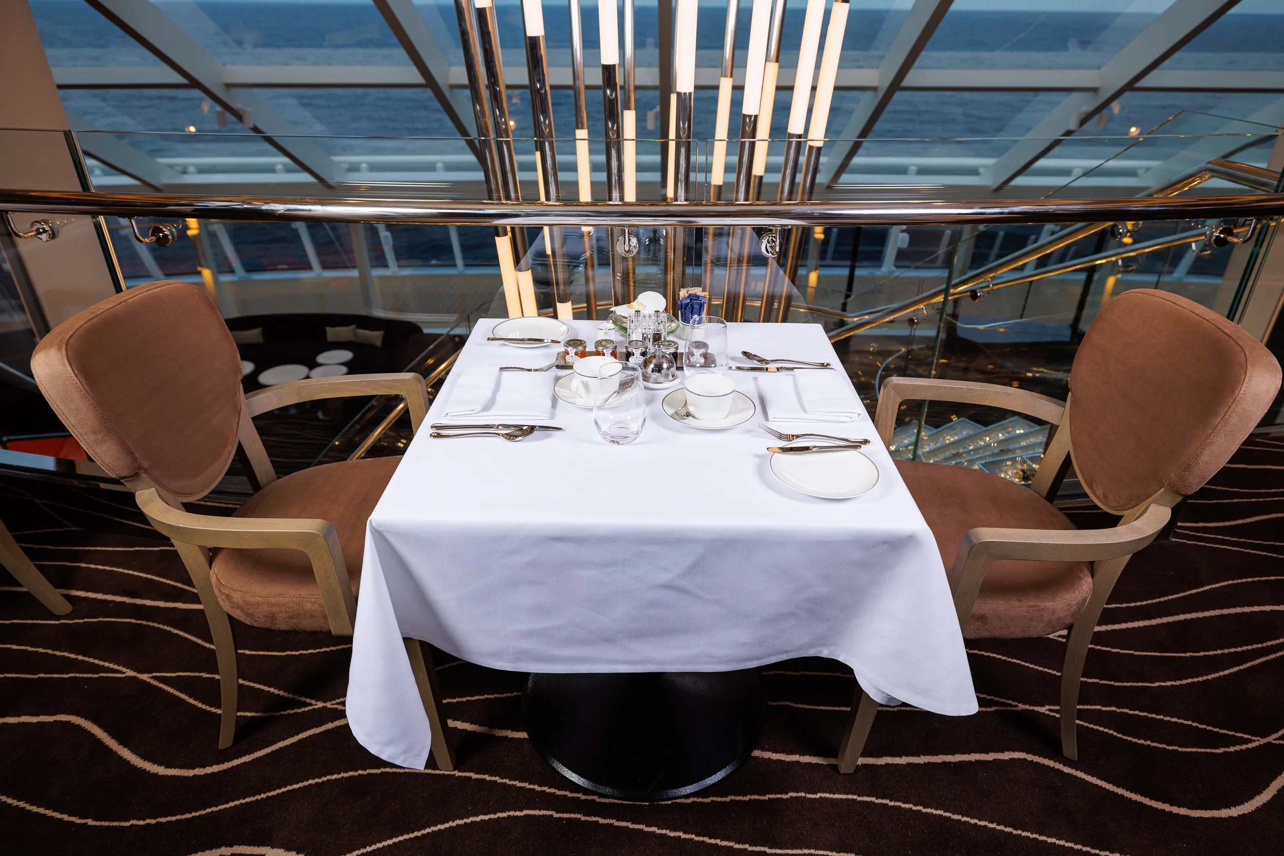 Table de restaurant élégante avec assiettes, verres, couverts en face de fenêtres donnant sur la mer, dans un intérieur moderne.