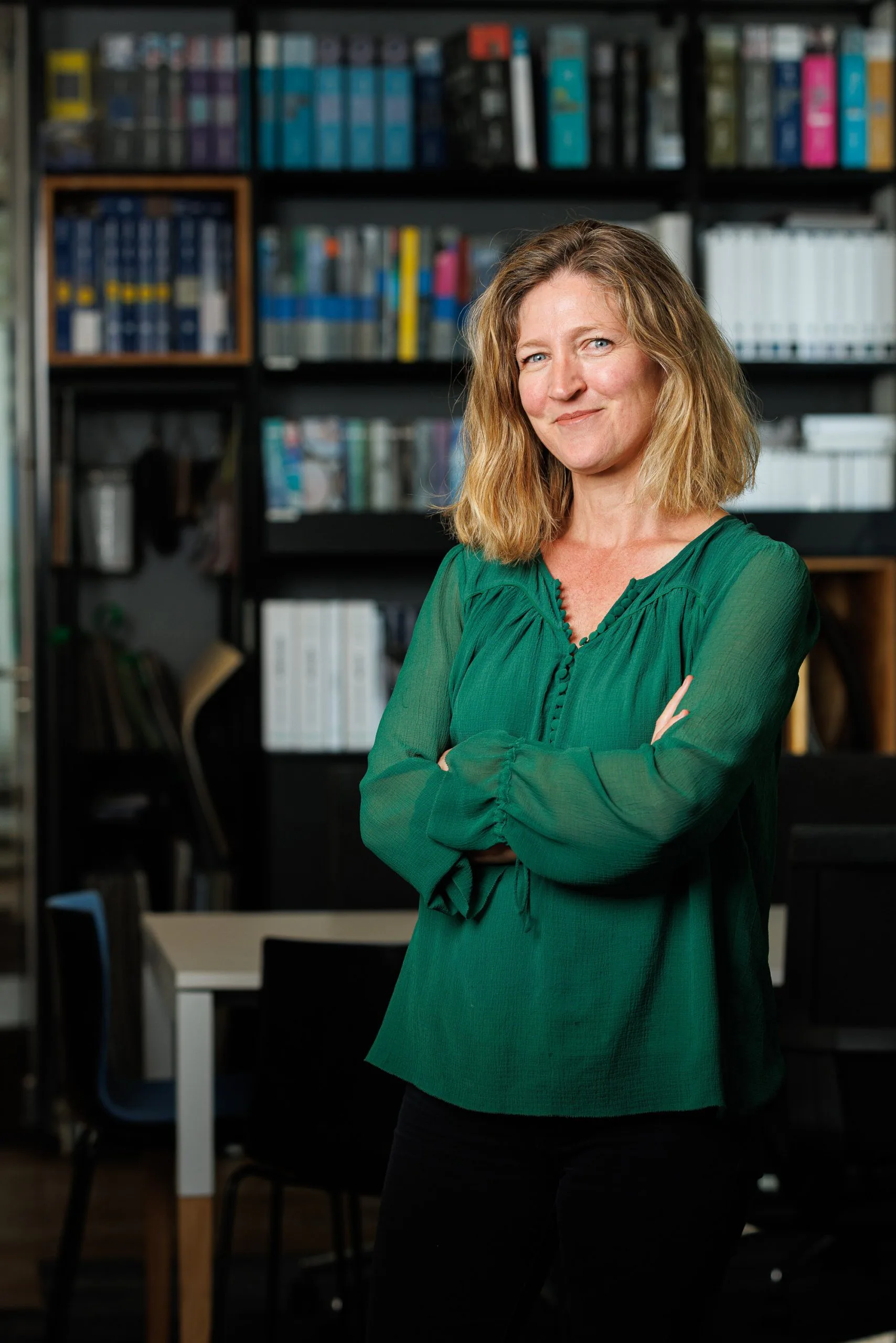 Femme souriante portant un chemisier vert devant une bibliothèque remplie de livres.
