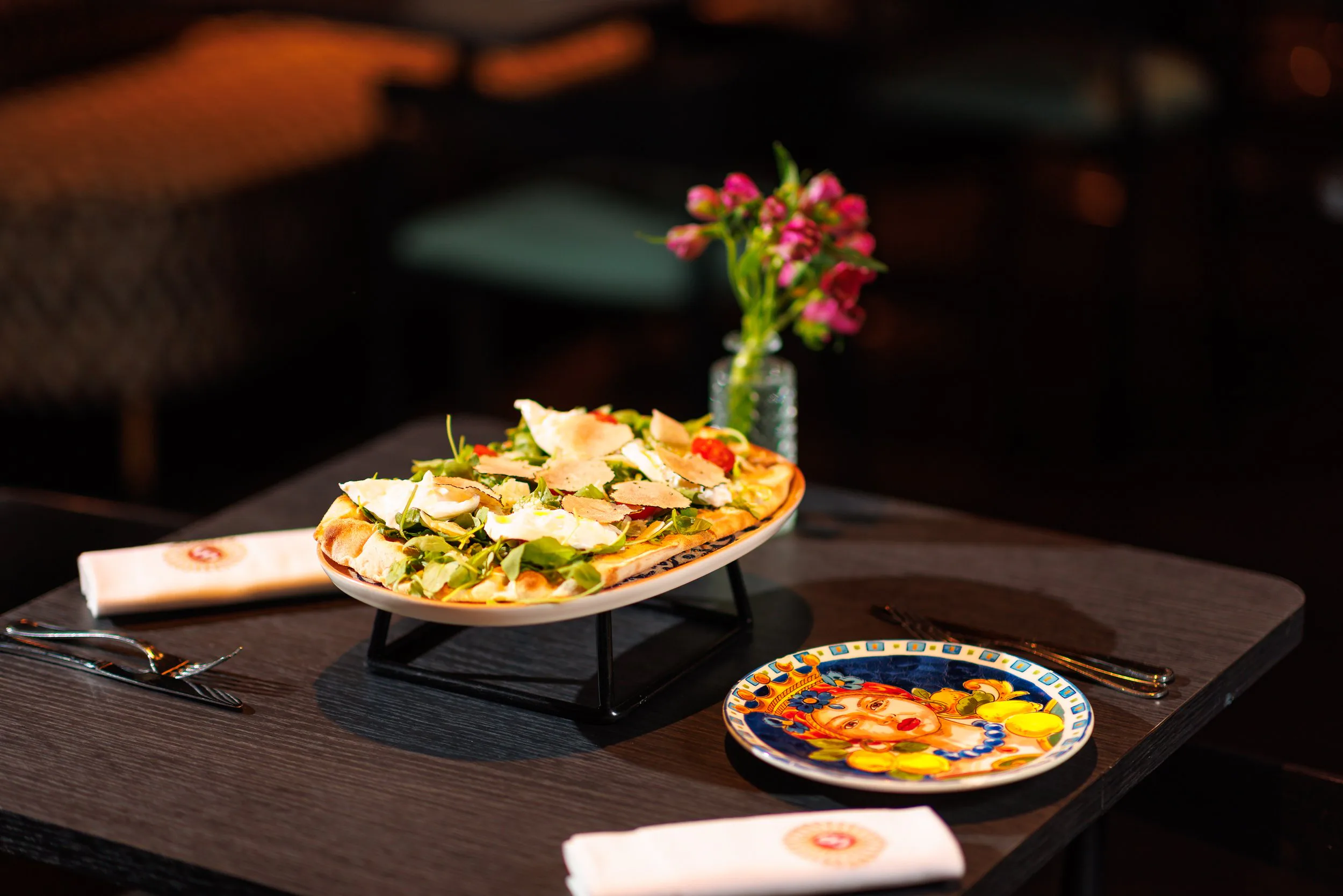 Une pizza garnie de légumes, servie sur un plat, accompagnée de couverts et d'une nappe blanche avec un motif rouge, sur une table en bois foncé. En arrière-plan, un vase avec des fleurs roses.