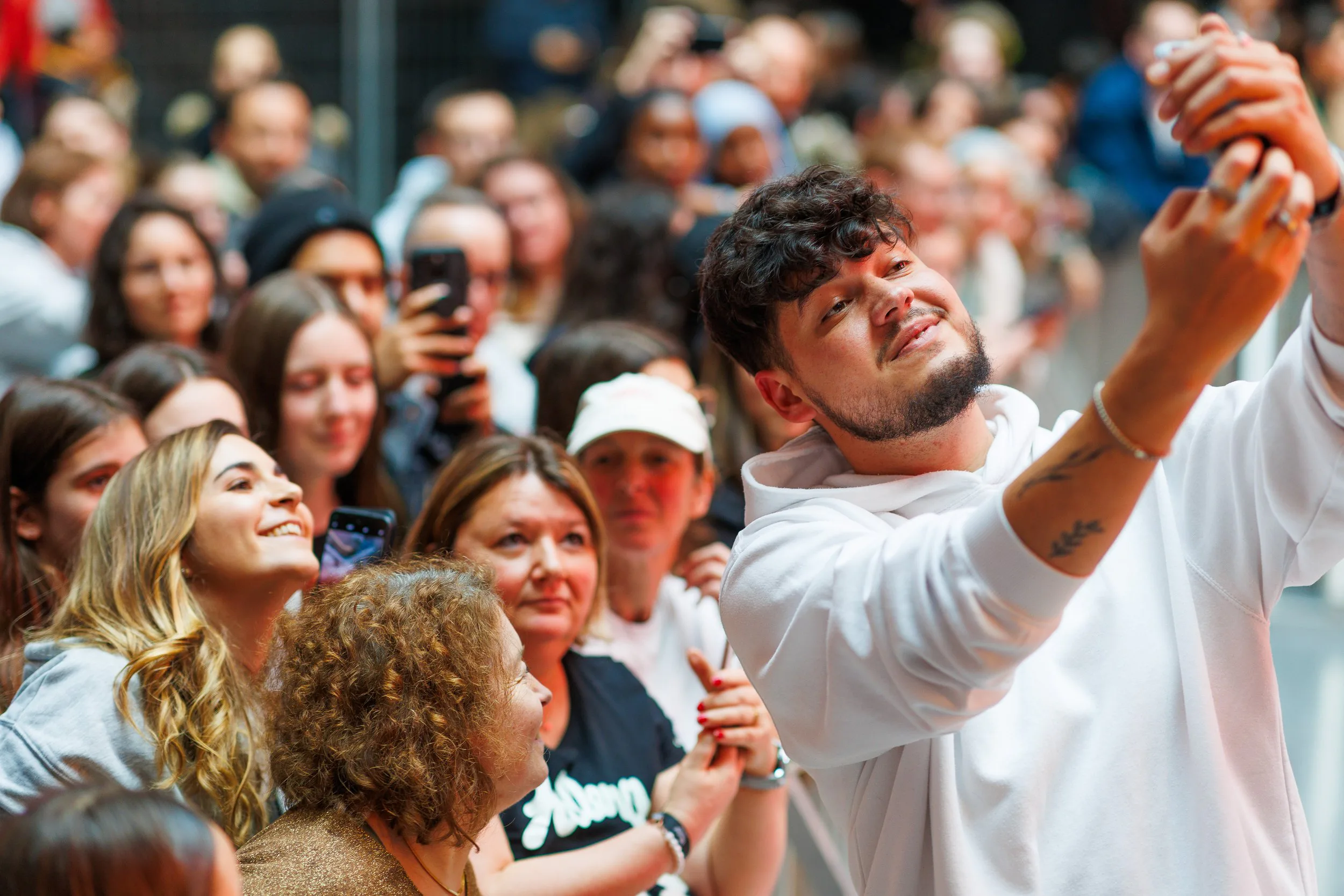 Groupe de personnes souriantes, dont un homme prenant une photo ou un selfie, lors d'un événement public. La scène est animée avec plusieurs spectateurs en arrière-plan.
