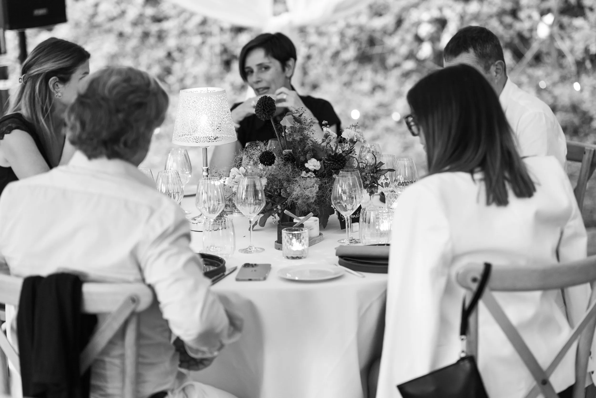 Groupe de personnes assises autour d'une table lors d'un dîner, avec une décoration florale centrale, une lampe, et une ambiance extérieure